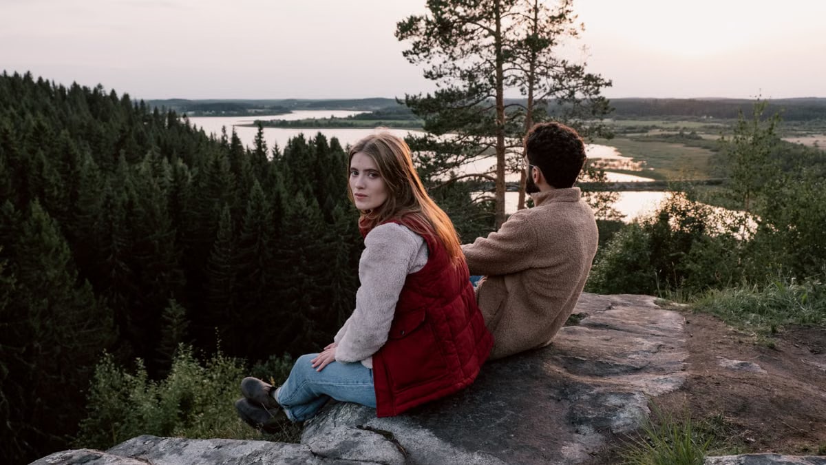 A couple sits on a rock overlooking a serene forest and river landscape at sunset.